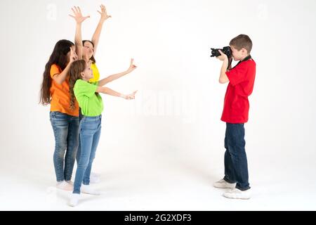 Ein junger Fotograf fotografiert im Studio drei niedliche Mädchen in hellen T-Shirts auf weißem Hintergrund. Junger Fotograf und Blogger. Schule o Stockfoto