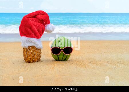 Bild von Ananas mit Weihnachtsmannhut und grüner, fröhlicher Wassermelone in Sonnenbrillen am tropischen Strand. Neujahrs- und Weihnachtskonzept. Stockfoto