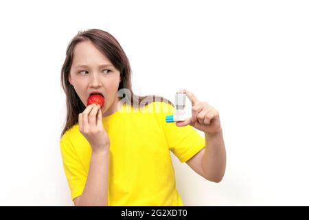 Ein 10-jähriges Mädchen hält Erdbeeren in den Händen und schaut vor Angst auf weißem Hintergrund auf den Asthmainhalator. Konzept für Lebensmittelallergien Stockfoto