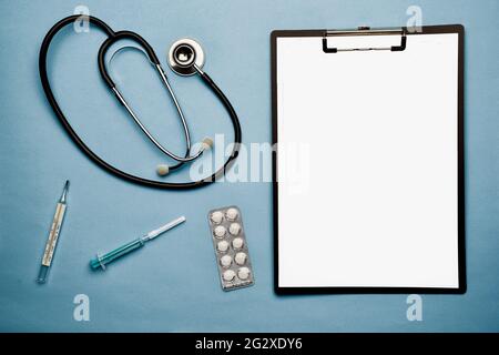 Auf blauem Hintergrund liegen ein Stethoskop, Pillen in einer Blisterpackung, eine Spritze, ein Thermometer und eine Notizmappe. Es gibt einen Platz für eine Inschrift Stockfoto