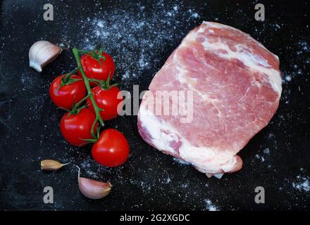Rohes Stück Schweinefleisch mit roten Tomaten, frischem Knoblauch und Salz auf schwarzem Hintergrund. Ansicht von oben auf ein Stück frisches Schweinefleisch ohne Knochen. Grill- und Food-Konzept. Stockfoto