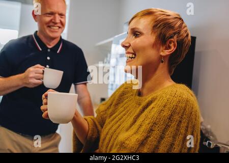 Reifer Mann und junge Frau stehen und halten Kaffeetasse im Büro. Fröhliche Führungskräfte stehen und machen eine Kaffeepause im Büro. Stockfoto