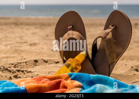 Den Helder, Niederlande, 3. Juni 2021. Nahaufnahme von Flip-Flops, Sonnencreme und einem farbenfrohen Handtuch am Strand von Huisduinen, Niederlande. Hochwertige Fotos. Nahaufnahme Stockfoto