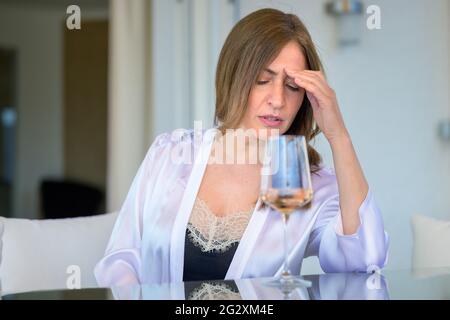 Introspective Frau sitzt tief in Gedanken an einem Tisch auf der Terrasse mit einem Glas Wein mit ihrer Hand auf der Stirn starrte beiseite mit einem Blick von Concentra Stockfoto