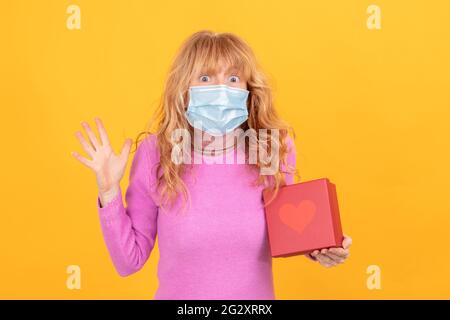 Erwachsene oder ältere Frau mit Gesichtsmaske isoliert auf dem Hintergrund Mit Geschenkbox mit Herz Stockfoto
