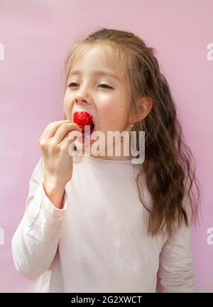 Ein glückliches niedliches Mädchen isst eine saftige süße Erdbeere auf einem rosa Hintergrund. Stockfoto