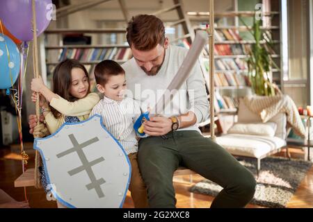 Ein glücklicher Vater sitzt auf der Schaukel in familiärer Atmosphäre zu Hause und spielt mit seinen Kindern. Familie, Freizeit, zusammen Stockfoto