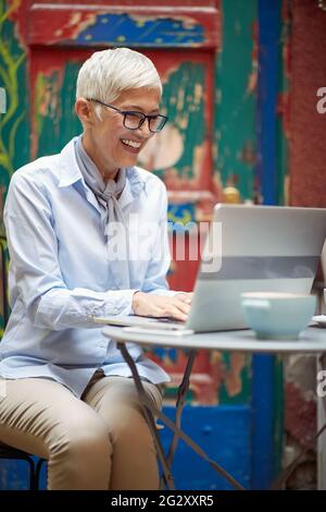 Schöne kaukasische ältere Frau, die im Café im Freien sitzt, auf ihrem Laptop tippt und lächelt Stockfoto