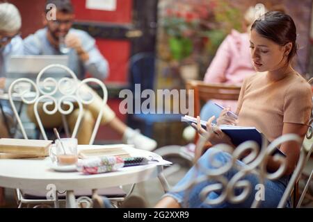 Junge Erwachsene kaukasische Frau, die im Café im Freien sitzt und allein ein Buch liest, mit anderen Menschen im Hintergrund. Stockfoto