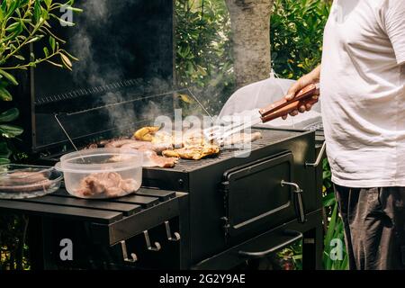 Mann beim Grillen von Fleisch auf einem Grill verschiedene Fleischsorten, Huhn, Würstchen, Blutwurst, Lamm auf einem Crbon-Grill im Garten Stockfoto