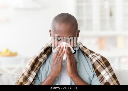 Kranker reifer afroamerikanischer Mann, der mit einer Decke bedeckt war, an Erkältung oder Allergien litt, seine Nase bläst und zu Hause sitzt Stockfoto