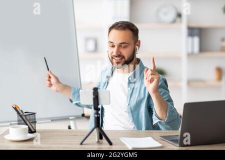 Glücklicher männlicher Lehrer, der online Privatunterricht von zu Hause aus gibt und auf eine leere Tafel mit leerem Platz zeigt Stockfoto