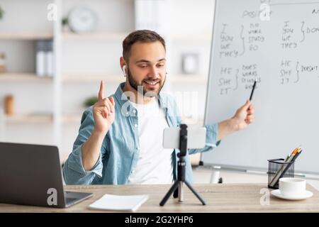 Junger männlicher Lehrer, der Online-Englischunterricht gibt, auf eine Tafel zeigt und die Grammatikregeln im Home Office erklärt Stockfoto