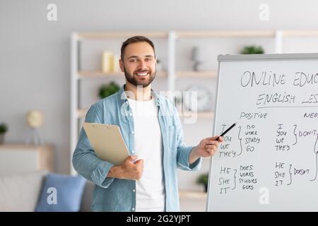 Fröhlicher junger männlicher Lehrer, der auf die Tafel zeigt, die Grammatik erklärt und Online-Englischunterricht vom Heimbüro gibt Stockfoto