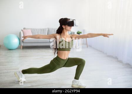 Virtuelles Training im Inland. Sportliche junge indische Dame in VR-Brille, die zu Hause im Krieger-Yoga-Asana steht Stockfoto