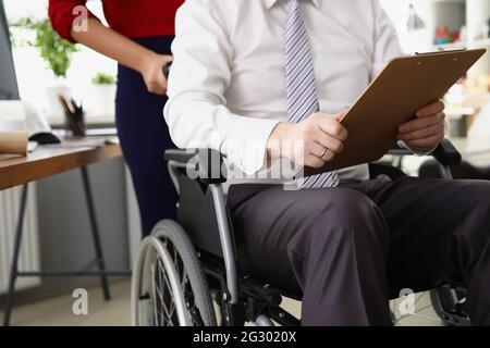Sekretärin rollt CEO im Rollstuhl mit Dokumenten und Berichten in den Händen Stockfoto
