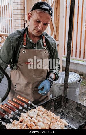 Grillparty im Hinterhof. Ein Mann mit Baseballmütze und Schürze bereitet für eine große Gruppe von Freunden einen Hähnchenfisch-Kebab vor. Street Food. Leckeres Fleisch Stockfoto