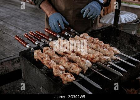 Der Koch bereitet Chicken Shish Kebab zu. Street Food. Fleisch über einem Feuer gekocht, Grillparty im Hinterhof. Männliche Hände in Handschuhen halten Stahlspieße mit d Stockfoto