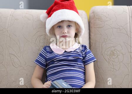 Kleines trauriges Mädchen, das auf dem Sofa sitzt und einen weihnachtsmannhut trägt Stockfoto