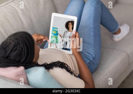 Unkenntlich schwarze Frau, die zu Hause auf dem Sofa liegt und sich den Online-Malkurs auf einem Tablet-Computer ansieht Stockfoto