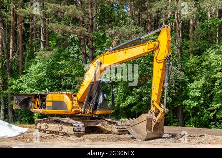 Der Bagger bereitet die Baustelle für den Bau der Straße vor. Gelbe Baumaschinen vor einem Hintergrund von üppig grünen Bäumen. Stockfoto