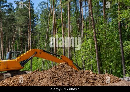 Der Bagger bereitet die Baustelle für den Bau der Straße vor. Orangefarbene Baumaschinen vor dem Hintergrund üppig grüner Bäume. Stockfoto