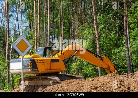 Der Bagger bereitet die Baustelle für den Bau der Straße vor. Orangefarbene Baumaschinen vor dem Hintergrund üppig grüner Bäume. Stockfoto