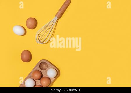 Eier in Holzkiste und Schneebesen auf gelbem Hintergrund. Flach liegend Draufsicht Kopierbereich. Stockfoto