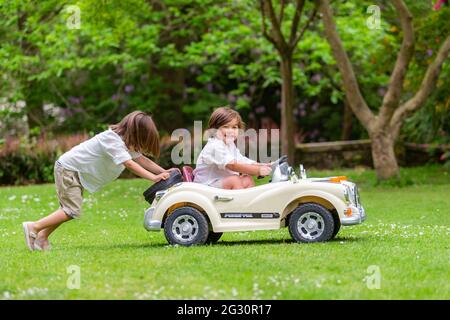 Zwei junge Brüder im Alter von sechs und drei Jahren spielen draußen mit einem Spielzeug-Elektroauto Stockfoto