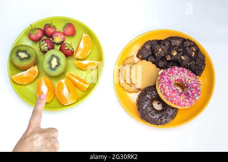 Gesund und ungesund, Obstteller vs Keksteller, Konzept, Finger zeigt gesunde Lebensmittel, heller Hintergrund Stockfoto