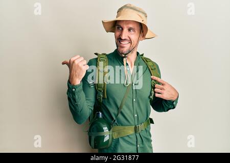 Hübscher Mann mit Bart, Explorer-Hut und Rucksack, lächelnd, glückliches Gesicht und mit Daumen nach oben zur Seite zeigend. Stockfoto