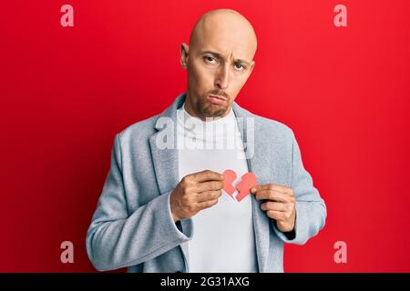 Kahlköpfiger Mann mit Bart, der gebrochene Herzpapierform hält, deprimiert und sorgt sich um Distress, weint wütend und ängstlich. Trauriger Ausdruck. Stockfoto