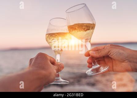 Verliebte Paare klirren mit Weißweingläsern, während sie den wunderschönen rosa Sonnenuntergang am Meer genießen. Romantischer Urlaub oder Dating-Konzept aus nächster Nähe im Stockfoto