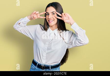 Junge Brünette Teenager tun Frieden Geste nahe den Augen lächeln mit einem glücklichen und kühlen Lächeln auf dem Gesicht. Zeigt Zähne. Stockfoto