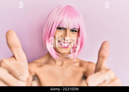 Junger Mann trägt Frau Make-up trägt rosa Perücke Genehmigung tun positive Geste mit der Hand, Daumen hoch lächelnd und glücklich für den Erfolg. Sieger Geste. Stockfoto