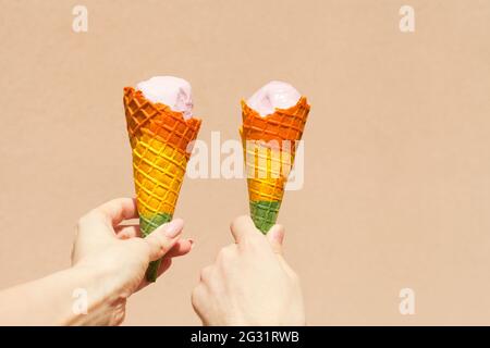 regenbogen-Eiskegel - zwei Hände halten Eiskegel in Regenbogenfarben an heißen Sommertagen nahe rosa Hintergrund Stockfoto