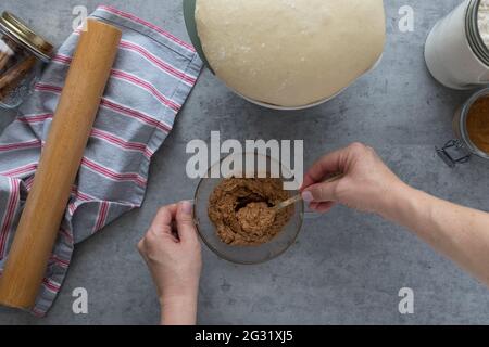 Frau mixt Zimtpulver, Panela und Butter mit einem Löffel in einer Kristallschüssel neben einem aufgestiegenen Teig. Stockfoto