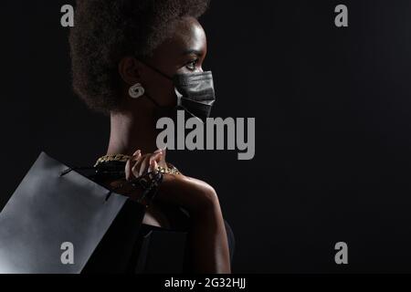 Profilansicht der afroamerikanischen Frau in schwarzer medizinischer Gesichtsmaske hält schwarze Einkaufstaschen. Safety on Black Friday Konzept. Covid-19 Coronavirus-Schutz. Stockfoto