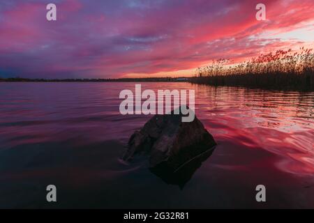Heller, wolkig kontrastierender, roter Sonnenuntergangshimmel, der sich im Wasser des Sees spiegelte, mit Stein auf der Vorderseite. Das Wasser hinunter Stockfoto