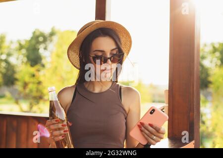 Junge schöne Frau in einem Sommerhut und Sonnenbrille, trinkt einen Drink und nutzt im Sommer im Park ein Mobiltelefon im Freien Stockfoto