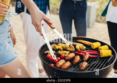 Mann macht Grill, verschiedene Gemüse und Hähnchenflügel mit Würstchen, Grillen auf einem tragbaren Grill im Freien in einem Park in der Natur Stockfoto