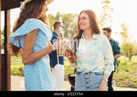 Zwei junge lächelnde junge Frauen, die an einem sonnigen Sommertag in Sommerkleidung und Hüten ein Picknick im Freien machen, kalte Getränke trinken Stockfoto