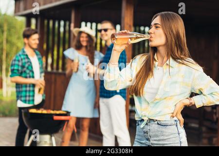 Attraktive junge Frau, mit einer Gruppe von Studenten Freunde beim Grillen, mit Biergetränken, eine Pause in der Natur an einem sonnigen Tag im Garten, picni Stockfoto