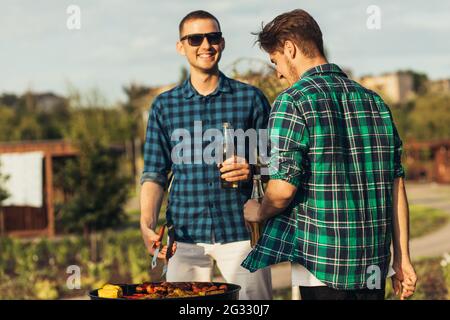 Zwei junge Männer mit Biergetränken, Grillen in der Natur, modische Menschen, die Fleisch zubereiten, Jugendstil, Essen, Freundschaft und Sommerkonzept Stockfoto