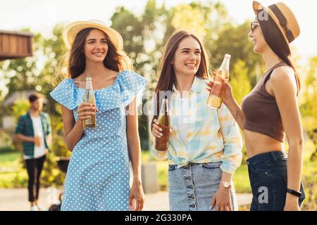 Drei schöne Freundinnen, die Spaß auf einem Picknick haben, Getränke trinken und plaudern, eine Gruppe junger Freunde, die auf dem Grill im Park grillen Stockfoto