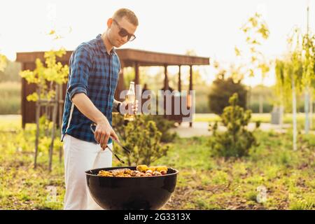 Mann macht Grill, verschiedene Gemüse und Hähnchenflügel mit Würstchen, Grillen auf einem tragbaren Grill im Freien in einem Park in der Natur Stockfoto