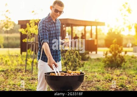 Mann macht Grill, verschiedene Gemüse und Hähnchenflügel mit Würstchen, Grillen auf einem tragbaren Grill im Freien in einem Park in der Natur Stockfoto