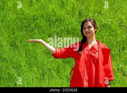 Ein lächelndes Mädchen in roten Kleidern auf einem Hintergrund aus grünem Gras, eine Handbewegung in der Seite. Eine junge, selbstbewusste Frau im Hintergrund des grünen Rasens hob ihre Hand zur Seite. Platz für Werbung Stockfoto