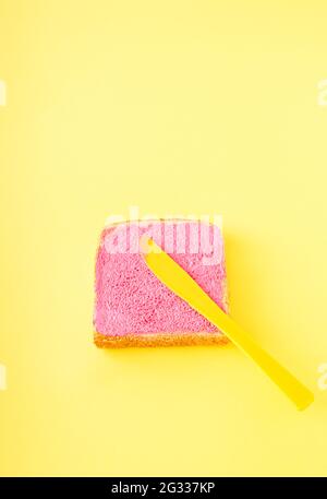 Rosa Neonfarbe auf einem Stück Brot auf einem gelben Hintergrund verschmiert. Das gelbe Messer ist auf dem Brot. Kreatives Food-Konzept, minimalistische Fotografie. Stockfoto