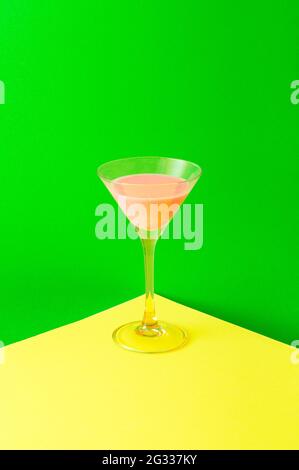 Ein Glas, das auf der gelben Ecke steht und mit einem pinken Neongetränk auf einem grünen Neonhintergrund gefüllt ist. Party minimalistisches Konzept mit lebendigen Farben. Stockfoto